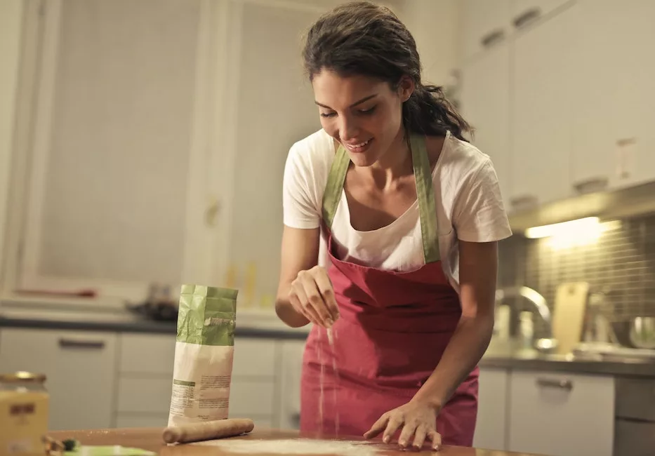 Low angle of positive young female in apron sprinkling flour over table while preparing food in mode